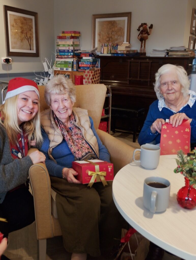2 elderly ladies sat in armchairs opening christmas presents and smiling with a carer