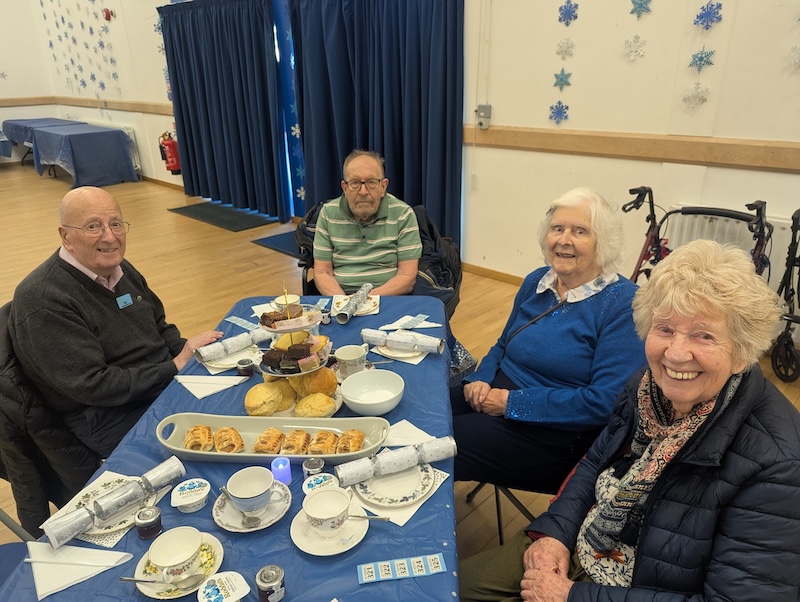 a group of elderly adults sat smiling together around a table in a community hall