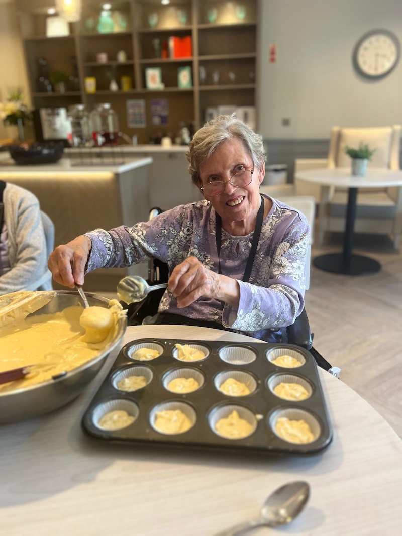elderly woman sat down smiling using spoons to put cupcake mixture into a baking tray