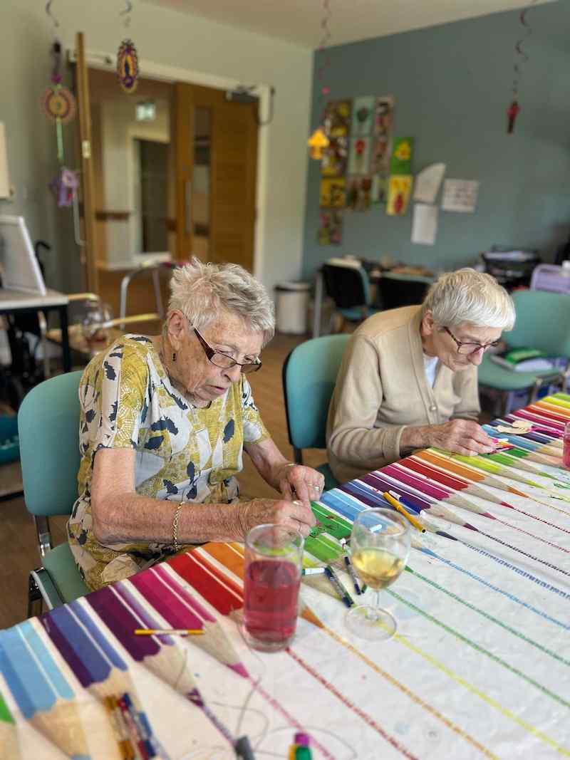 2 female residents sat down at a table with a colouring pencil tablecloth on doing colouring