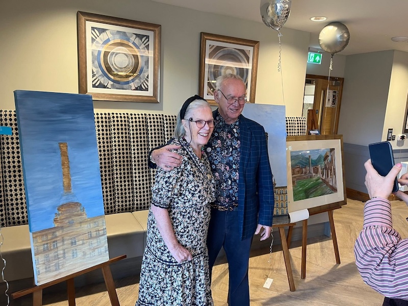2 of our residents stood together smiling in front of a showcase of paintings